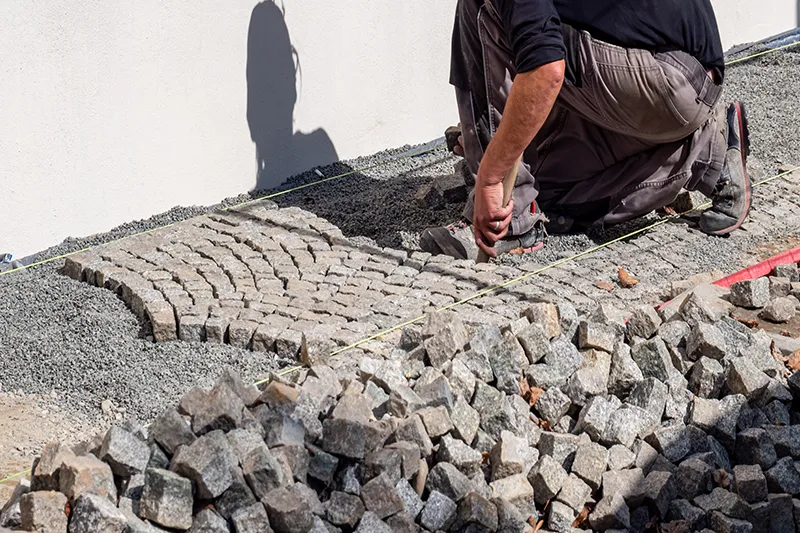 Eine Person verlegt Pflastersteine ​​auf einer Kiesfläche neben einer weißen Wand. Die Szene vermittelt ein Gefühl von Handarbeit und Handwerkskunst.