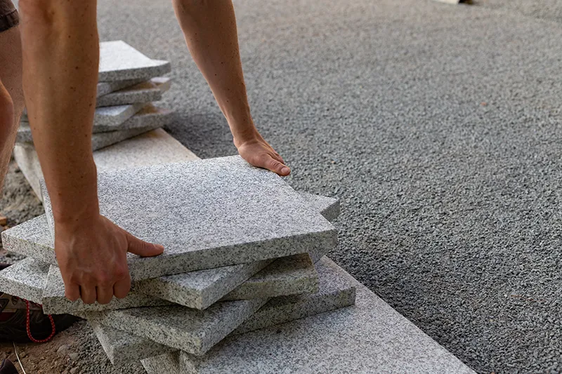 Eine Person arrangiert rechteckige Steinplatten auf einer Kiesfläche und legt dabei Wert auf sorgfältige Platzierung. Die Szene vermittelt Präzision und Handarbeit bei der Landschaftsgestaltung.